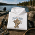 A white pullover hoodie is folded on a wooden dock with a lake and forest in the background. The hoodie has a graphic print of Bigfoot holding a fish with the text 'GONE TILL Further Notice.
