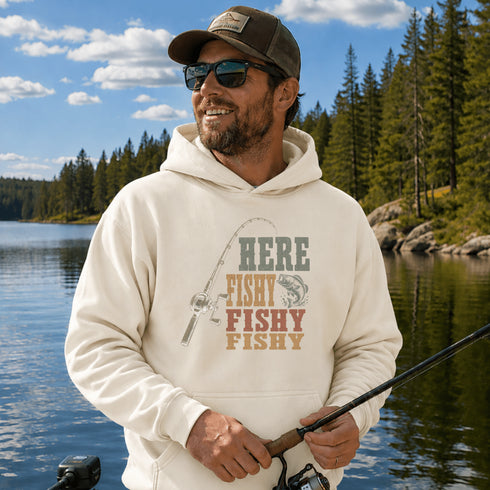 A man wearing a cream-colored hoodie with a fishing graphic and the text 'HERE FISHY FISHY FISHY' stands by a lake with trees in the background, holding a fishing rod.