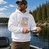 Person wearing a unisex hoodie with a vintage-inspired fishing graphic while holding a fish on a boat surrounded by a scenic lake and forest. Suitable for lifestyle, outdoor, fishing, cabin, camping, and product mockups.