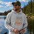 Person wearing a unisex hoodie with a vintage-inspired fishing graphic while holding a fish on a boat surrounded by a scenic lake and forest. Suitable for lifestyle, outdoor, fishing, cabin, camping, and product mockups.
