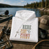 A folded white hoodie with a rustic fishing-themed graphic reading 'HERE FISHY FISHY FISHY' is displayed on a wooden dock by a lake, surrounded by a fishing rod, tackle box, cap, mug, and fishing net.