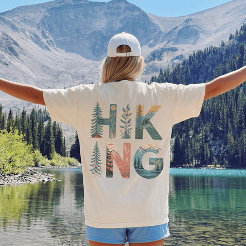 Person wearing a 'Hiking' shirt with nature-themed design, standing by a lake with mountains in the background.