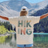 Person wearing a 'Hiking' shirt with nature-themed design, standing by a lake with mountains in the background.
