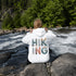woman wearing hiking hoodie with large nature graphic on back sitting on a rock at the lake