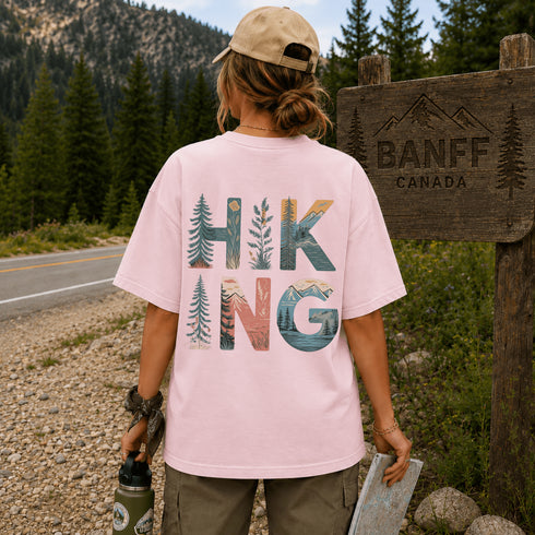 Person wearing a pink t-shirt with 'Hiking' design, standing near a Banff Canada sign.