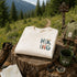 White sweatshirt with 'Hiking' design on a wooden stump in a forest setting.
