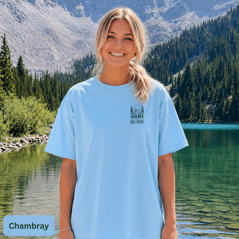 Home Is Out Here Comfort Colors 1717 graphic t-shirt featuring mountain, pine trees, and wilderness artwork with a small left chest logo and large back print.