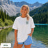 Home Is Out Here Comfort Colors 1717 graphic t-shirt featuring mountain, pine trees, and wilderness artwork with a small left chest logo and large back print.