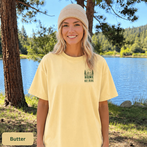 Home Is Out Here Comfort Colors 1717 graphic t-shirt featuring mountain, pine trees, and wilderness artwork with a small left chest logo and large back print.