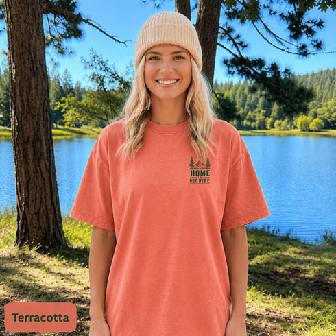 Home Is Out Here Comfort Colors 1717 graphic t-shirt featuring mountain, pine trees, and wilderness artwork with a small left chest logo and large back print.