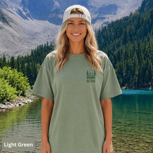 Home Is Out Here Comfort Colors 1717 graphic t-shirt featuring mountain, pine trees, and wilderness artwork with a small left chest logo and large back print.