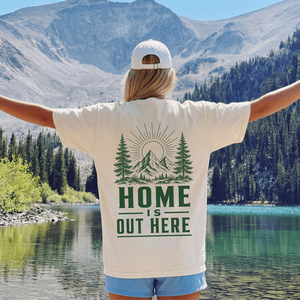 Home Is Out Here Comfort Colors 1717 graphic t-shirt featuring mountain, pine trees, and wilderness artwork with a small left chest logo and large back print.
