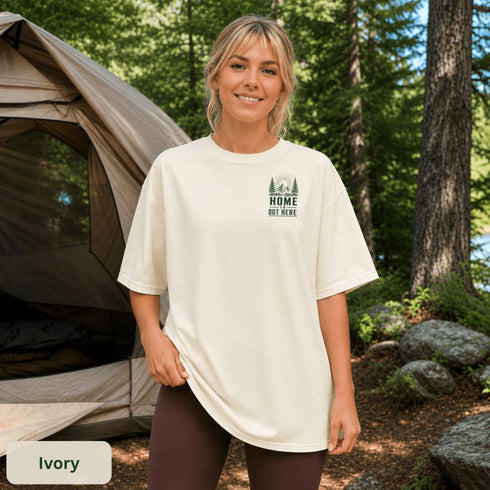 Home Is Out Here Comfort Colors 1717 graphic t-shirt featuring mountain, pine trees, and wilderness artwork with a small left chest logo and large back print.