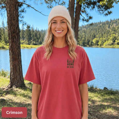 Home Is Out Here Comfort Colors 1717 graphic t-shirt featuring mountain, pine trees, and wilderness artwork with a small left chest logo and large back print.