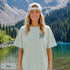 Home Is Out Here Comfort Colors 1717 graphic t-shirt featuring mountain, pine trees, and wilderness artwork with a small left chest logo and large back print.