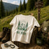 Home Is Out Here Comfort Colors 1717 graphic t-shirt featuring mountain, pine trees, and wilderness artwork with a small left chest logo and large back print.