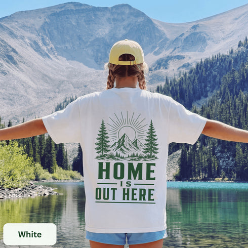 Home Is Out Here Comfort Colors 1717 graphic t-shirt featuring mountain, pine trees, and wilderness artwork with a small left chest logo and large back print.