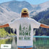 Home Is Out Here Comfort Colors 1717 graphic t-shirt featuring mountain, pine trees, and wilderness artwork with a small left chest logo and large back print.
