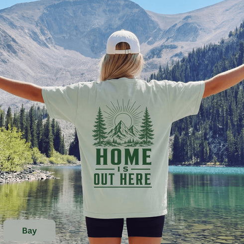 Home Is Out Here Comfort Colors 1717 graphic t-shirt featuring mountain, pine trees, and wilderness artwork with a small left chest logo and large back print.