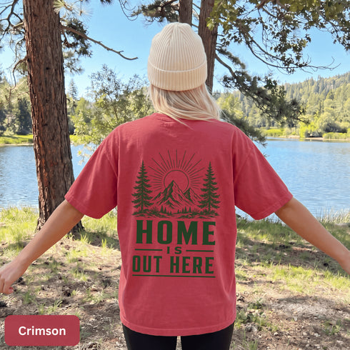 Home Is Out Here Comfort Colors 1717 graphic t-shirt featuring mountain, pine trees, and wilderness artwork with a small left chest logo and large back print.