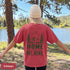 Home Is Out Here Comfort Colors 1717 graphic t-shirt featuring mountain, pine trees, and wilderness artwork with a small left chest logo and large back print.