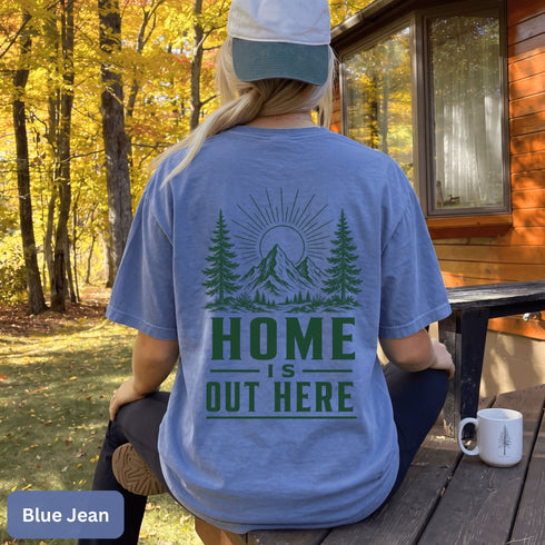 Home Is Out Here Comfort Colors 1717 graphic t-shirt featuring mountain, pine trees, and wilderness artwork with a small left chest logo and large back print.
