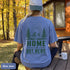 Home Is Out Here Comfort Colors 1717 graphic t-shirt featuring mountain, pine trees, and wilderness artwork with a small left chest logo and large back print.