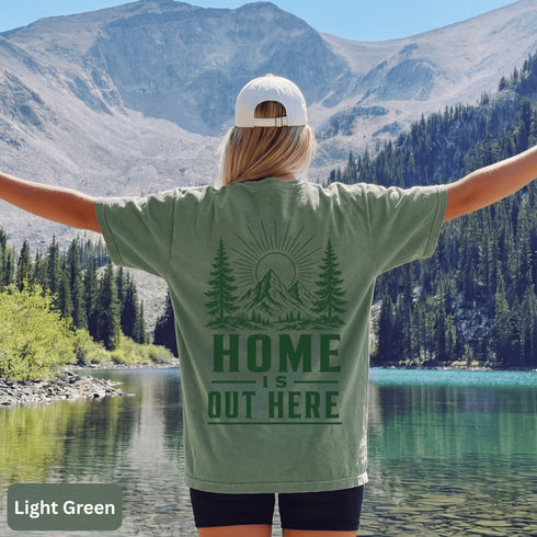 Home Is Out Here Comfort Colors 1717 graphic t-shirt featuring mountain, pine trees, and wilderness artwork with a small left chest logo and large back print.