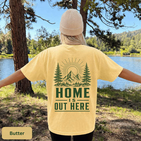 Home Is Out Here Comfort Colors 1717 graphic t-shirt featuring mountain, pine trees, and wilderness artwork with a small left chest logo and large back print.