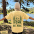 Home Is Out Here Comfort Colors 1717 graphic t-shirt featuring mountain, pine trees, and wilderness artwork with a small left chest logo and large back print.