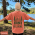 Home Is Out Here Comfort Colors 1717 graphic t-shirt featuring mountain, pine trees, and wilderness artwork with a small left chest logo and large back print.