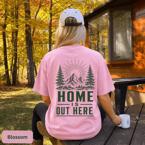 Home Is Out Here Comfort Colors 1717 graphic t-shirt featuring mountain, pine trees, and wilderness artwork with a small left chest logo and large back print.