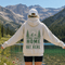 A person standing outdoors in front of a mountain lake, wearing a cream hoodie with a green forest and mountain graphic and the text 'HOME IS OUT HERE' on the back, along with a white cap and dark shorts.