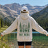 A person standing outdoors in front of a mountain lake, wearing a cream hoodie with a green forest and mountain graphic and the text 'HOME IS OUT HERE' on the back, along with a white cap and dark shorts.