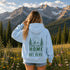 A woman stands in a mountain meadow with pine trees and snow-capped peaks in the background, viewed from behind. She is wearing a light blue hoodie with a dark green graphic on the back showing mountains, trees, and the text 'HOME IS OUT HERE'.