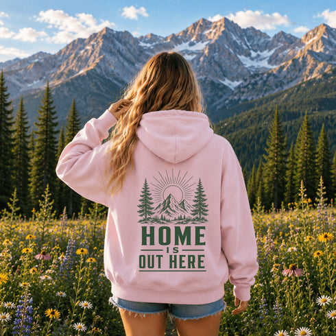 A woman stands with her back to the camera in a mountain meadow, wearing a light pink hoodie with a green forest and mountain graphic on the back that reads 'HOME IS OUT HERE'.