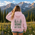 A woman stands with her back to the camera in a mountain meadow, wearing a light pink hoodie with a green forest and mountain graphic on the back that reads 'HOME IS OUT HERE'.