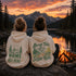 Two people sitting on a rock by a campfire at sunset, seen from behind, wearing matching beige hoodies with green outdoor graphics and text. Mountains, a lake, and pine trees are visible in the background.