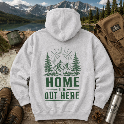 A light gray hooded sweatshirt laid flat on a rustic wooden surface, featuring a green graphic of mountains, pine trees, and the text 'HOME IS OUT HERE.' The hoodie is styled with outdoor props including boots, a backpack, a mug, a water bottle, a map, and a mountain lake background.