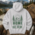 A light gray hooded sweatshirt laid flat on a rustic wooden surface, featuring a green graphic of mountains, pine trees, and the text 'HOME IS OUT HERE.' The hoodie is styled with outdoor props including boots, a backpack, a mug, a water bottle, a map, and a mountain lake background.