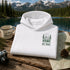 A folded white hoodie with a green 'HOME IS OUT HERE' forest graphic is displayed on a rustic wooden table. The scene is styled with a vintage coffee mug, a camera, a pinecone, and a map, with a scenic mountain lake backdrop.