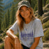 Woman sitting outdoors in a forest wearing a t-shirt with a nature-themed design.