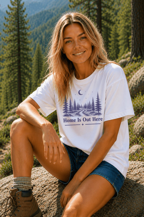 Woman sitting outdoors in a forest wearing a white t-shirt with a nature-themed design.