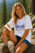 Woman sitting outdoors in a forest wearing a white t-shirt with a nature-themed design.