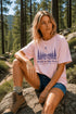 Woman sitting on a rock in a forest wearing a pink t-shirt with nature-themed design.