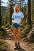 Woman standing on a forest path wearing a light blue t-shirt with a nature graphic and text.