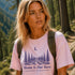 Woman wearing a pink t-shirt with nature design and 'Home Is Out Here' text in a forest setting
