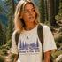 Woman hiking in a forest wearing a t-shirt with 'Home Is Out Here' and tree design.