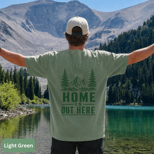 Home Is Out Here Comfort Colors 1717 graphic t-shirt featuring mountain, pine trees, and wilderness artwork with a small left chest logo and large back print.