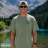 Home Is Out Here Comfort Colors 1717 graphic t-shirt featuring mountain, pine trees, and wilderness artwork with a small left chest logo and large back print.